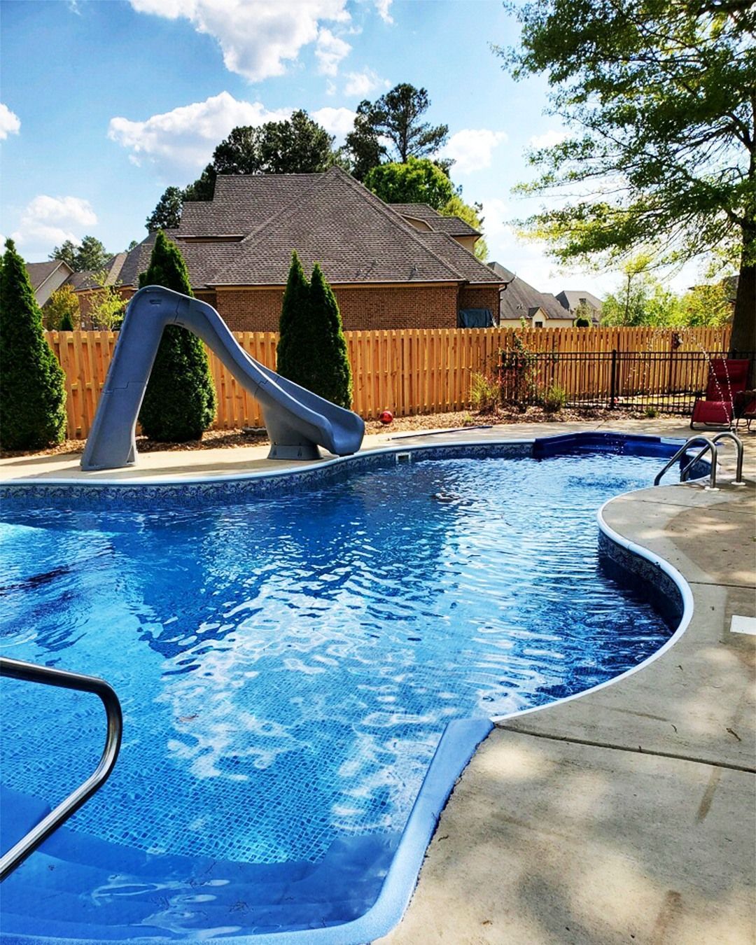 Backyard pool with blue water, slide, and wooden fence on a sunny day