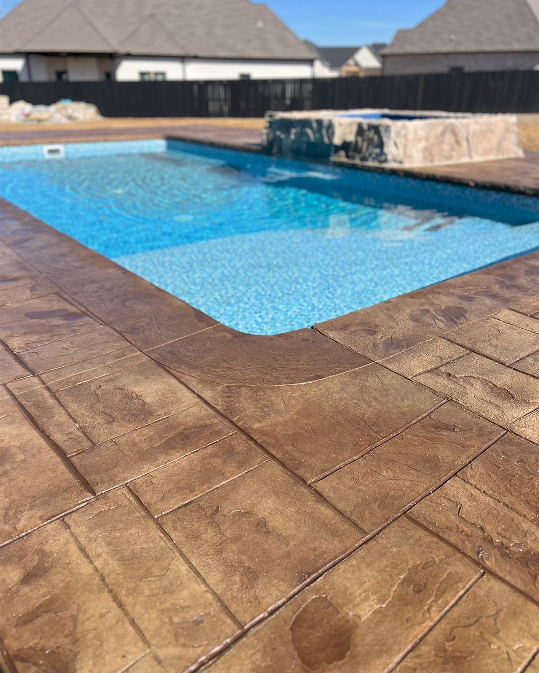 Blue swimming pool beside tan stone patio, with patio homes in the background.