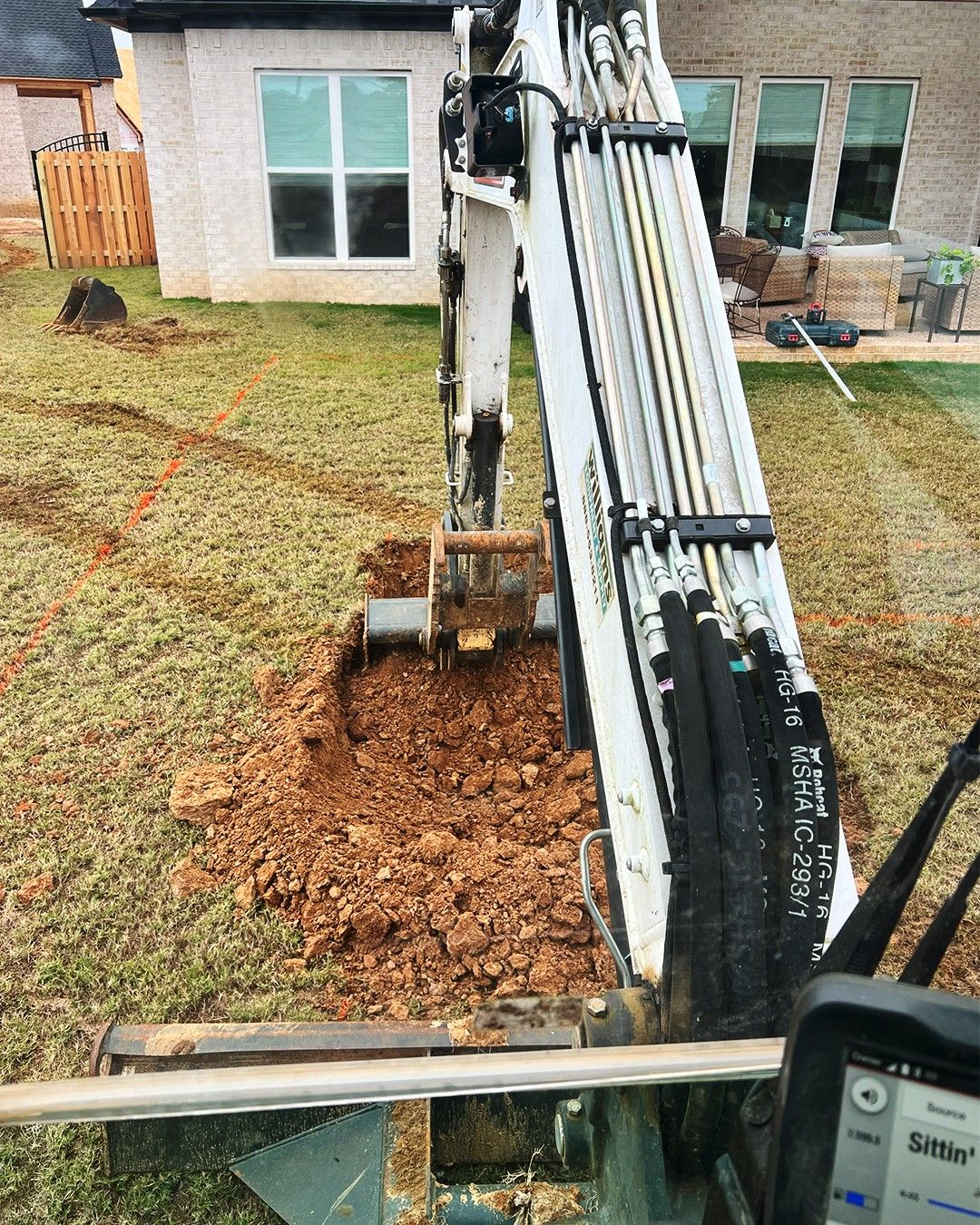 Excavator arm clearing leaves in a backyard near a house and patio furniture