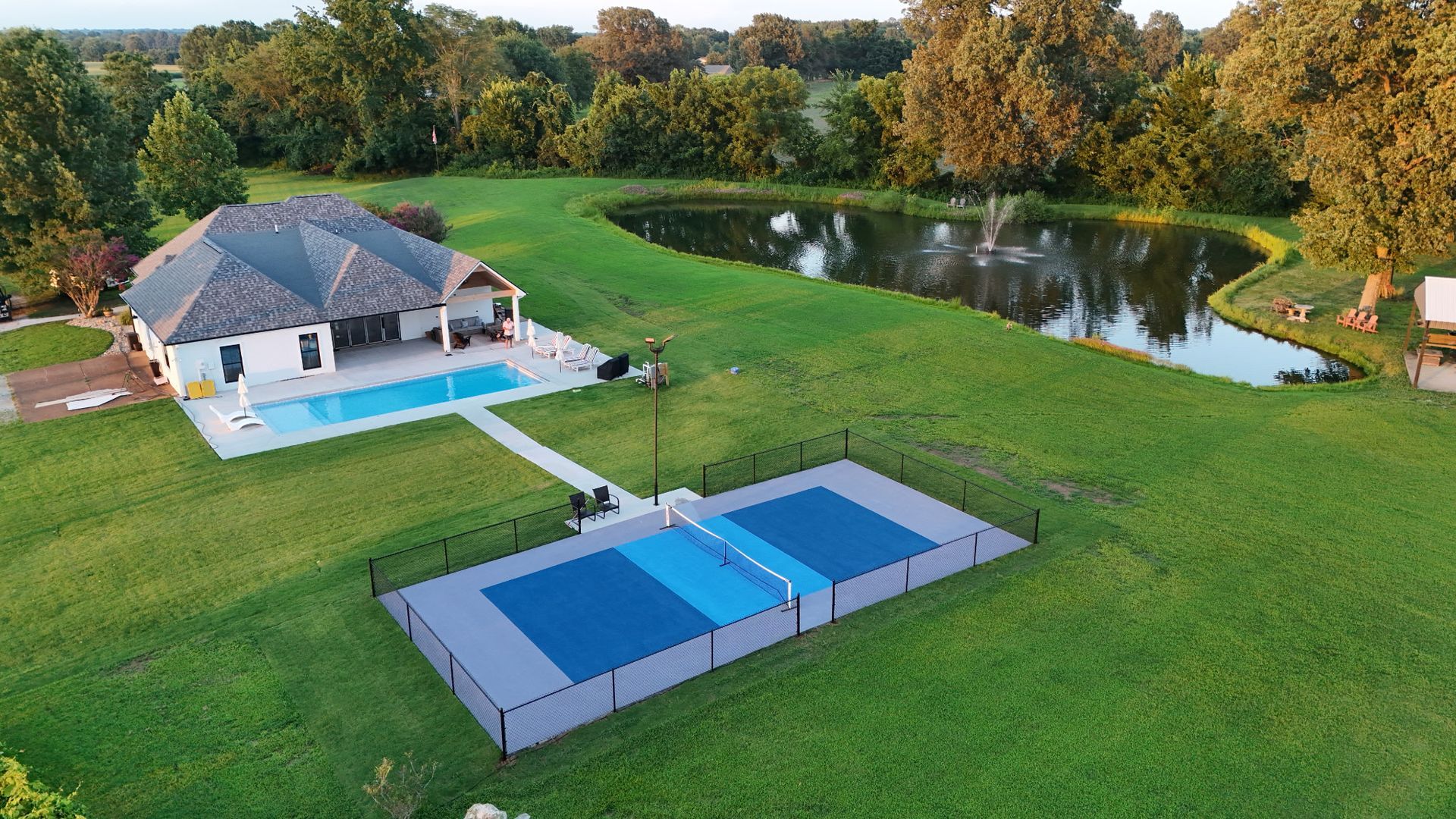 Backyard home with pool, tennis court, and pond surrounded by green lawn and trees