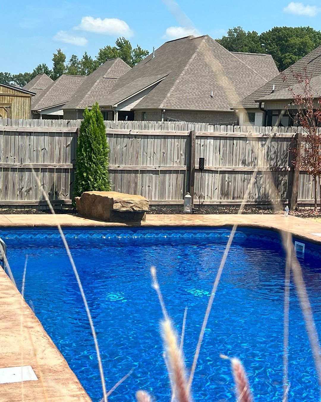 Backyard swimming pool with fountain jets, wooden fence, and neighboring rooftops under blue sky
