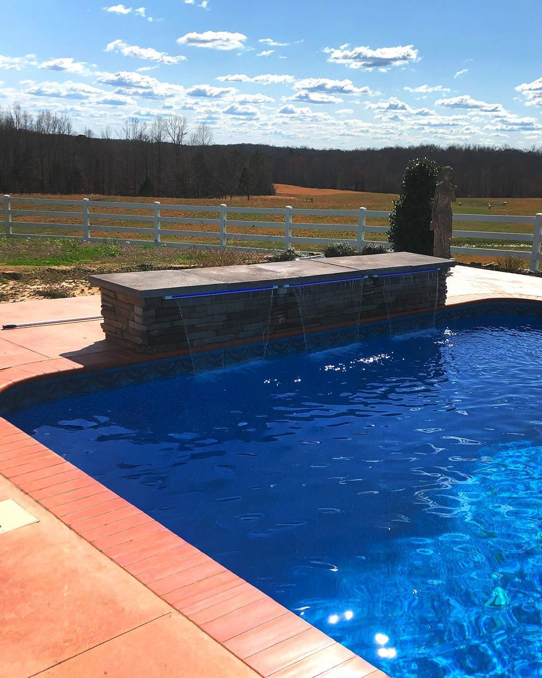 Backyard pool with blue water, stone ledge, and a field under a cloudy sky