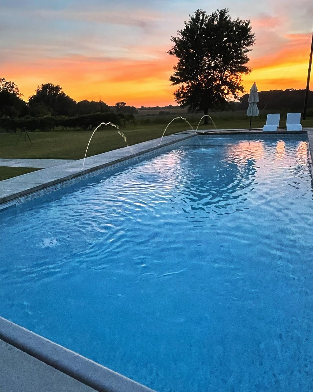 Backyard swimming pool at sunset with water jets, lounge chairs, and an orange sky