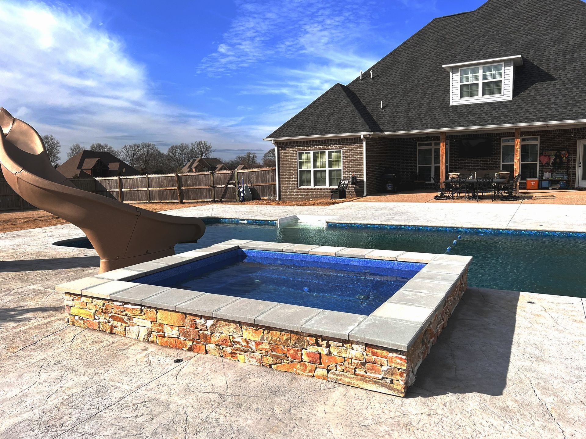Backyard pool with a stone hot tub, slide, and house under a blue sky