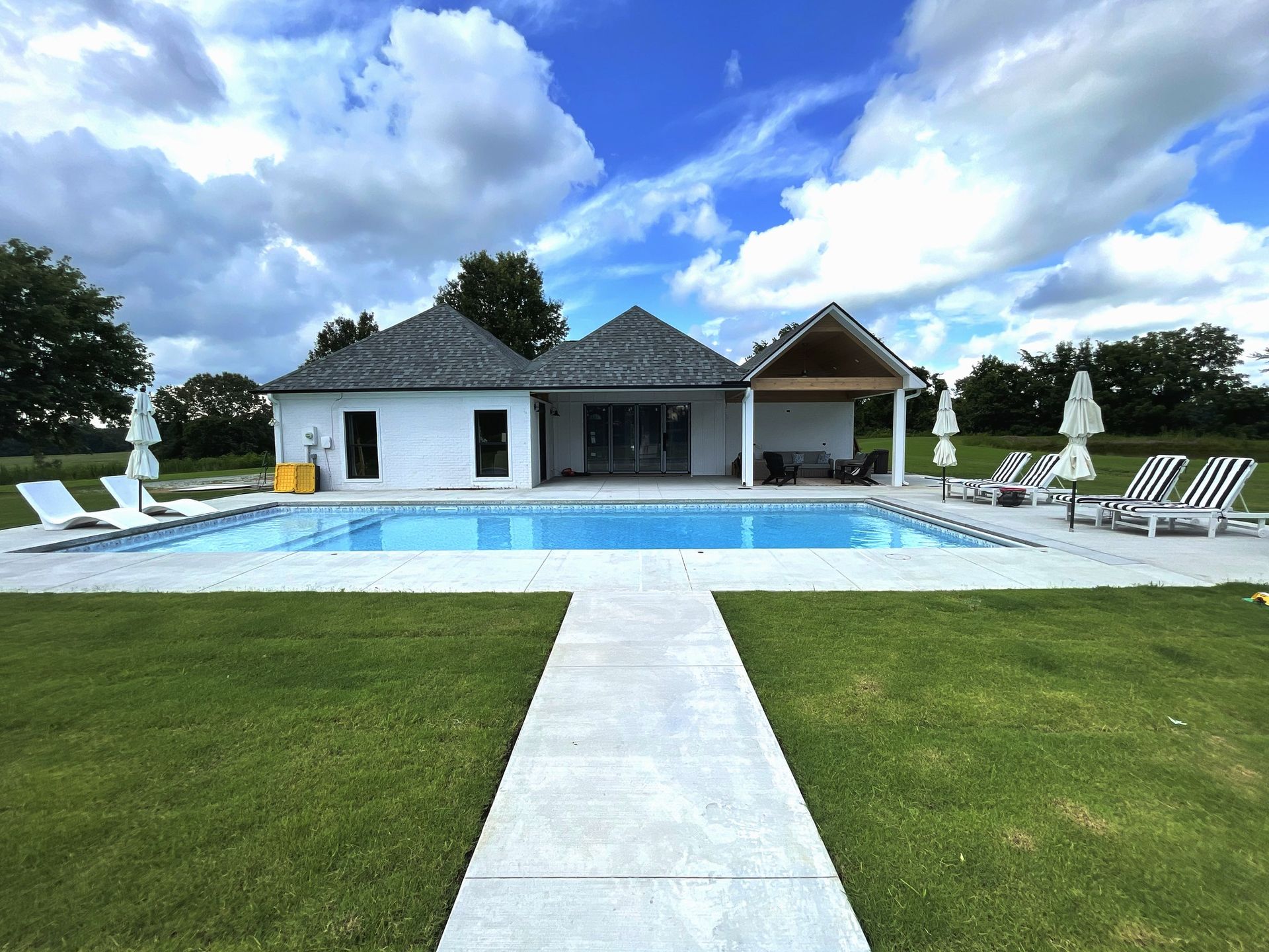 Modern house with a pool, white lounge chairs, and a green lawn under a cloudy sky