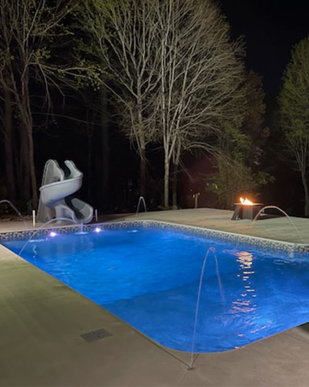 Lit backyard pool at night with a waterslide, glowing blue water, and bare trees nearby