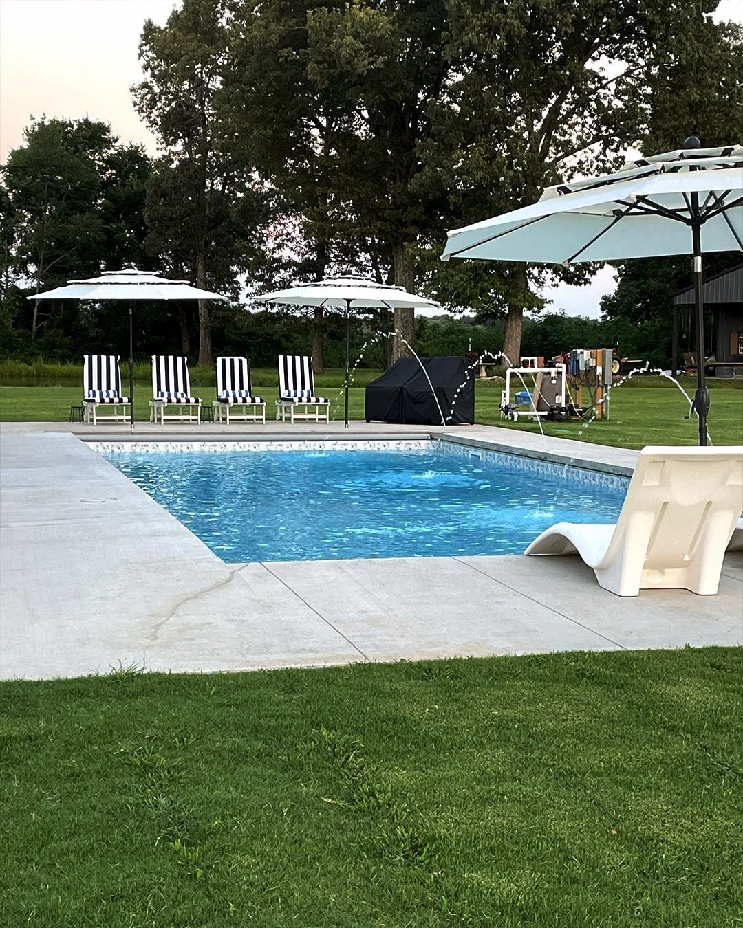 Backyard pool with lounge chairs, umbrellas, and green lawn at dusk