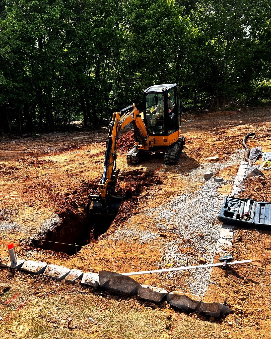 Small excavator digging a trench at a wooded construction site, with pipes laid nearby.
