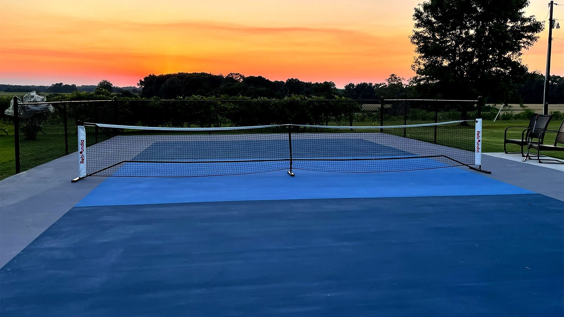 Outdoor tennis court at sunset, with a blue surface and net under an orange sky.