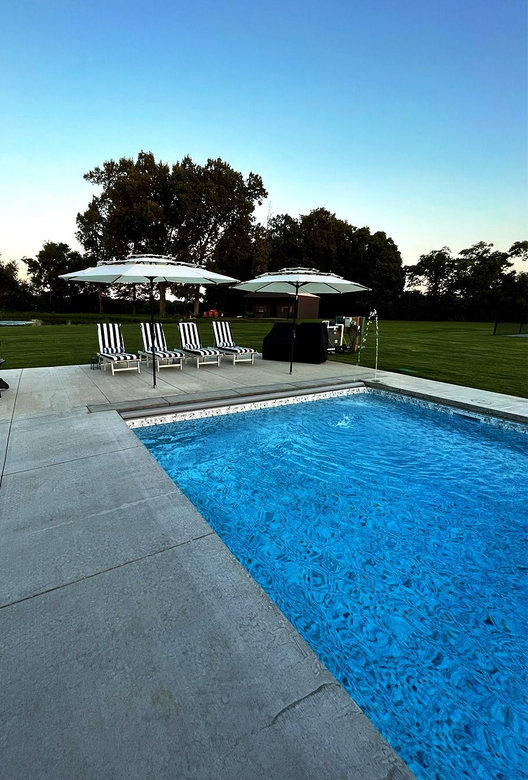Backyard swimming pool with patio chairs, grill, and trees at dusk