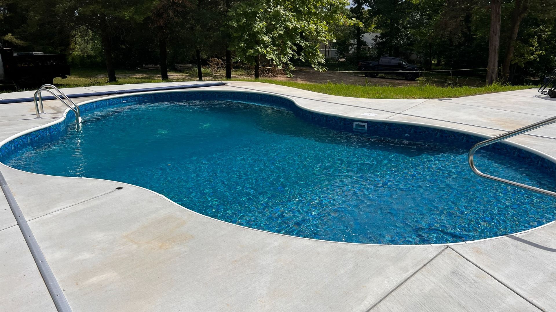 Backyard swimming pool with blue water, concrete deck, and trees in the background