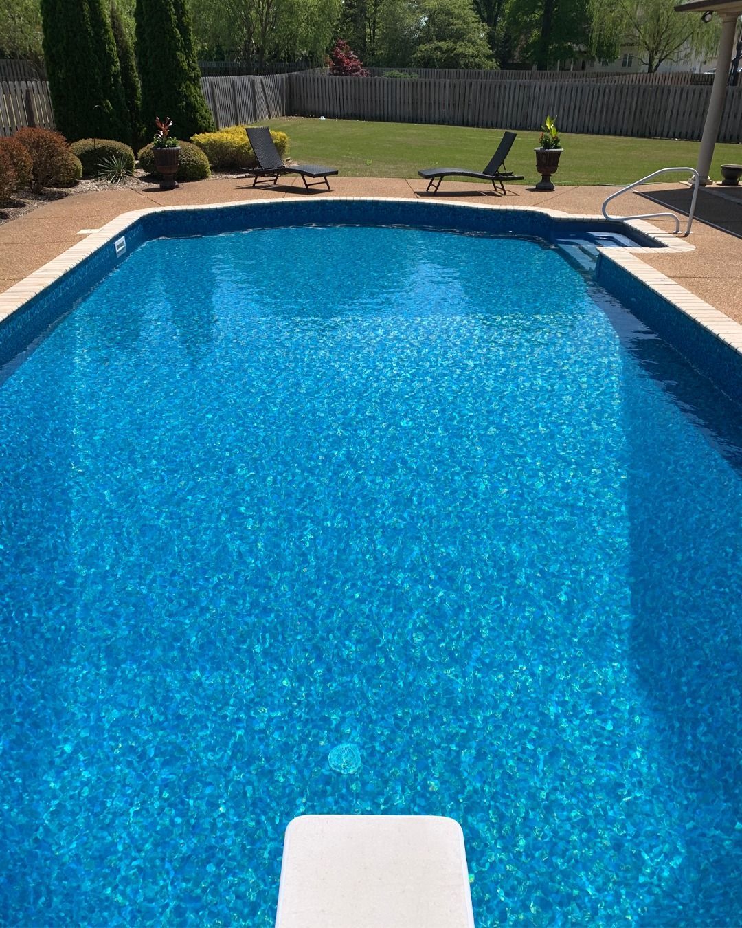 Blue swimming pool with a diving board and patio chairs in a sunny backyard