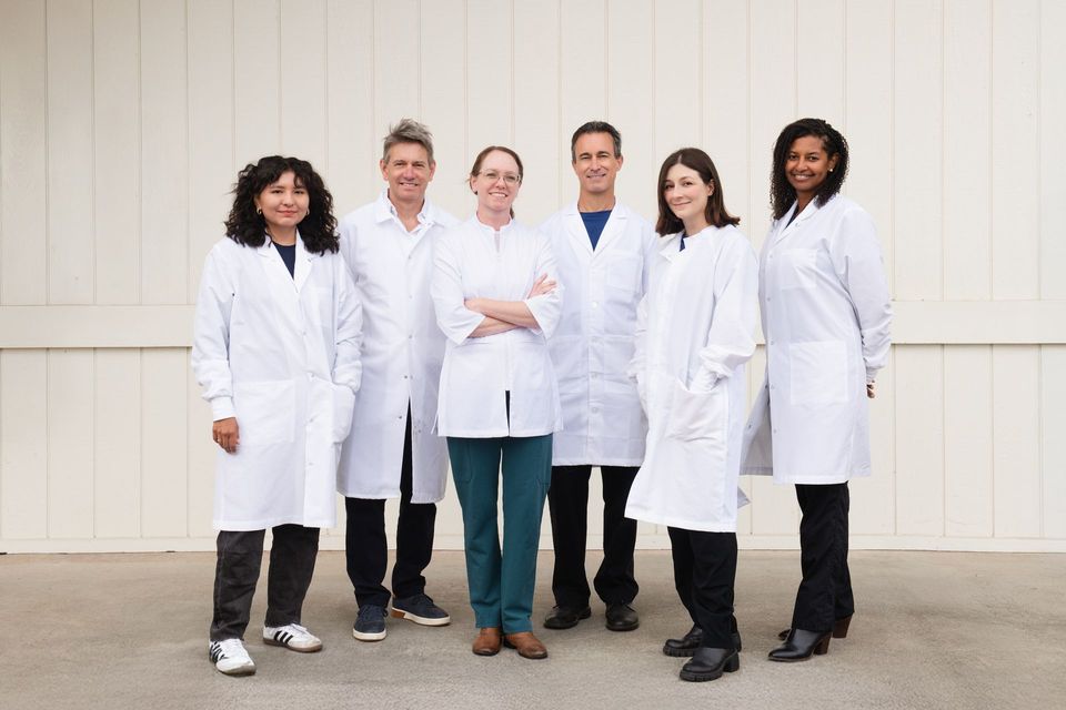 `Five people in white lab coats standing together against a light wall outdoors