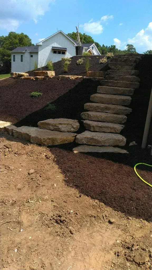 Stacked stone steps leading up a mulched embankment toward a white house on a sunny day.