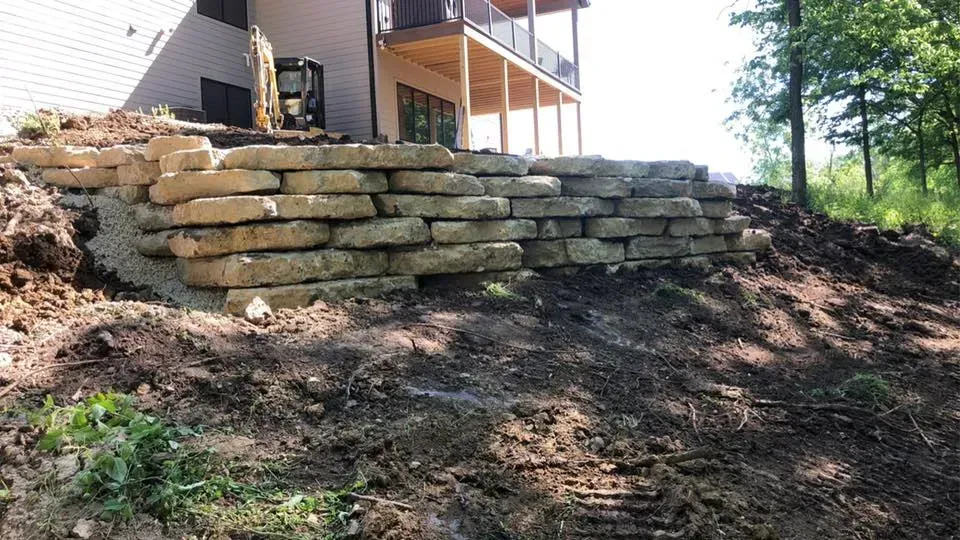 A tiered stone retaining wall under construction next to a house with a wooden deck and surrounding landscaping.