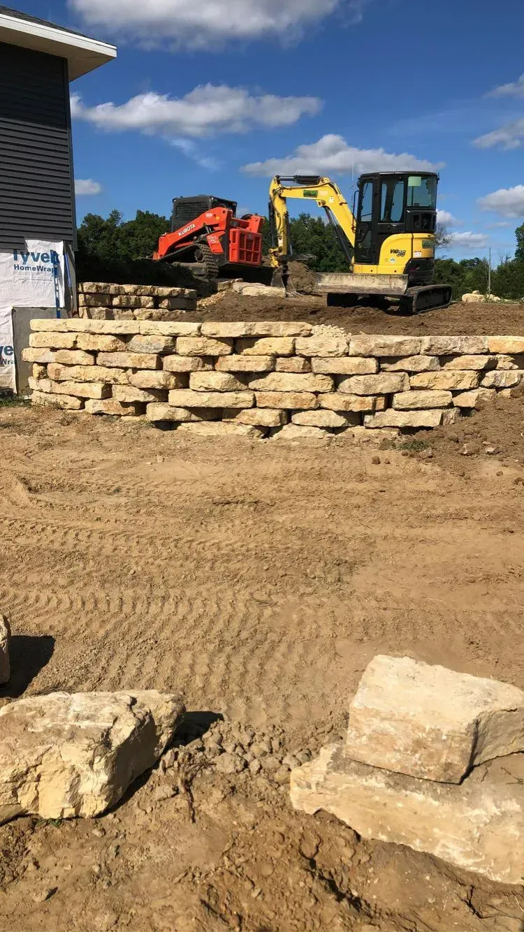 Construction site with an orange skid steer and yellow excavator behind a stacked stone retaining wall under a blue sky.