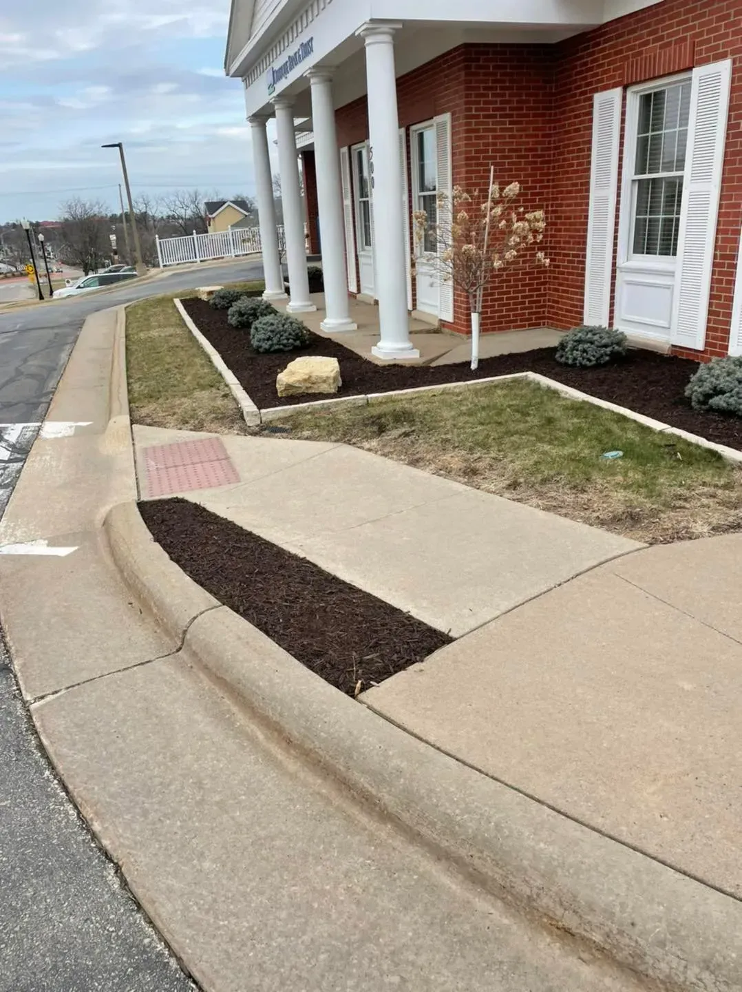Concrete sidewalk corner near a red brick building with white columns and manicured landscaping beds.