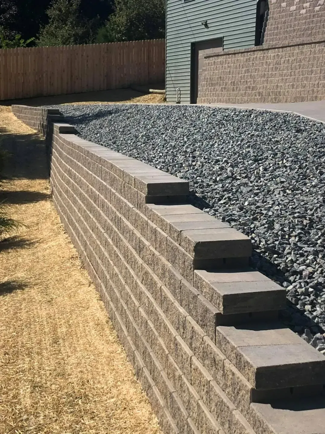 A tiered segmental retaining wall made of grey stone blocks, with a gravel bed behind it leading to a building.