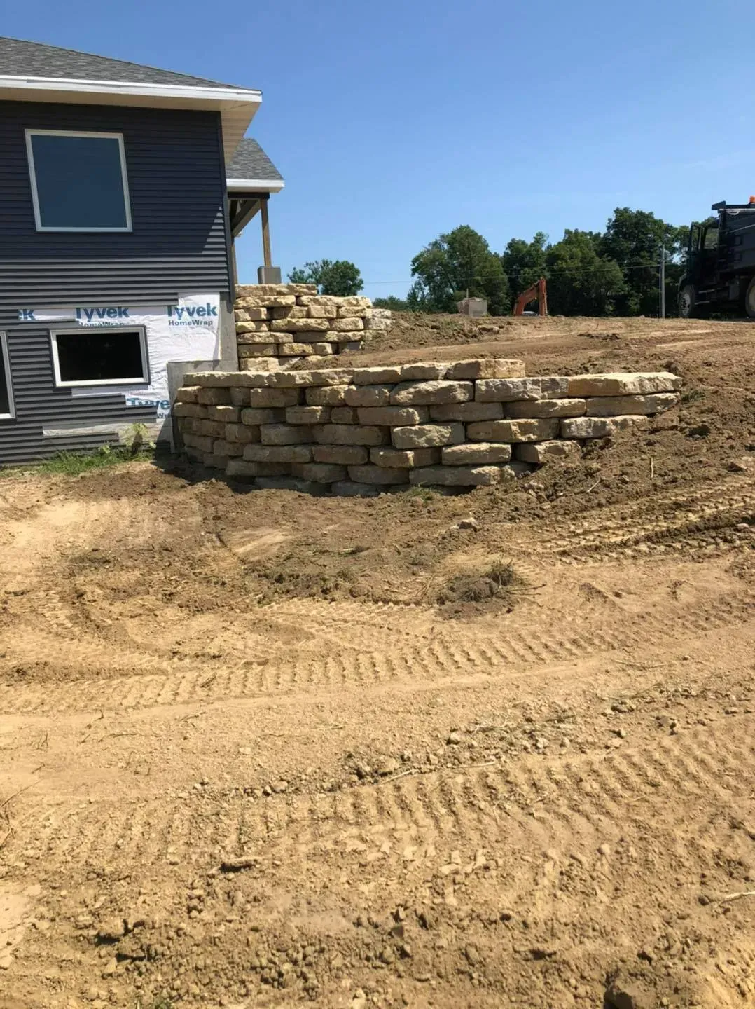 A newly built stone retaining wall stands against the side of a house under construction on a dirt lot.