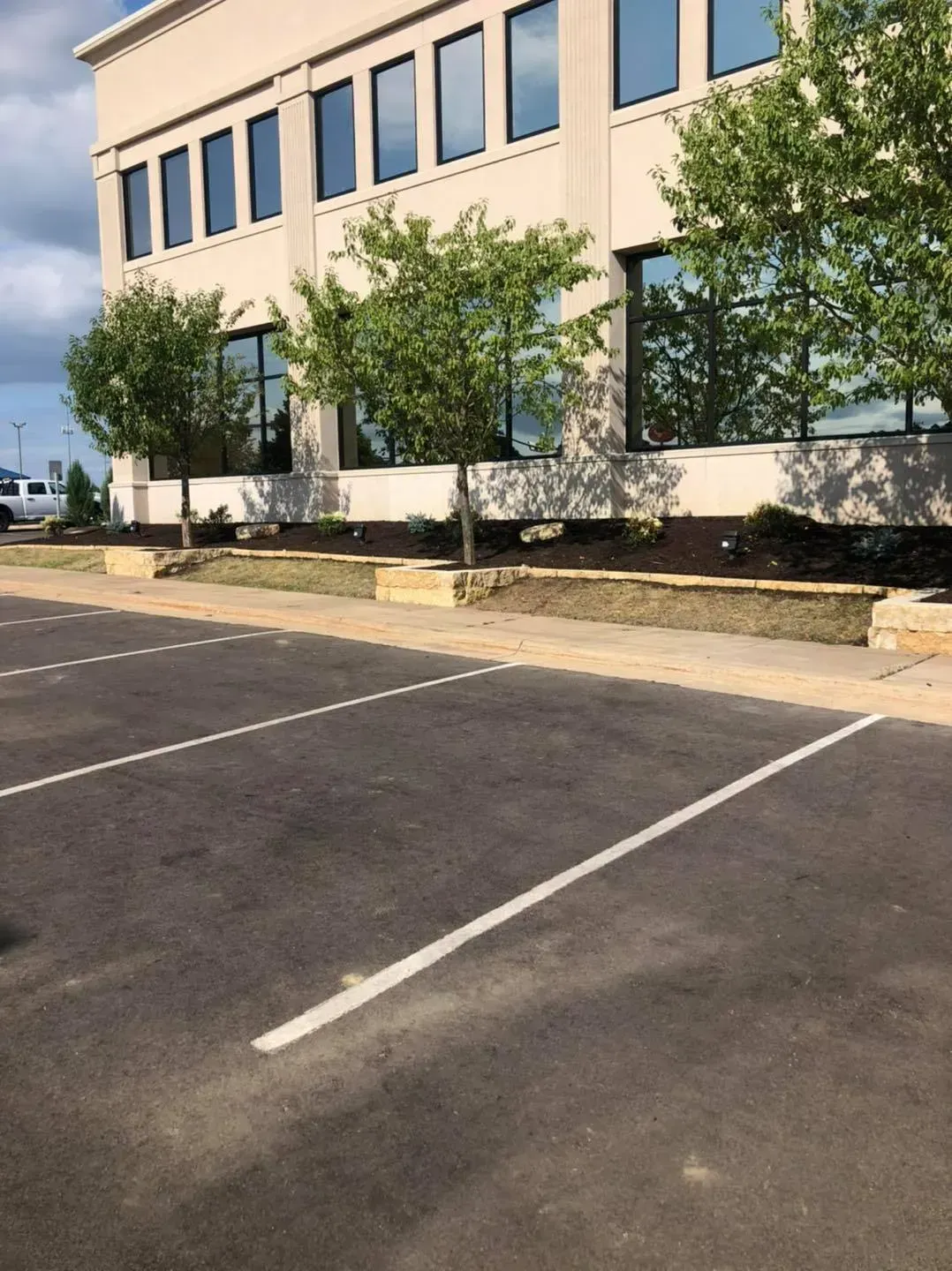 An asphalt parking lot in the foreground with a modern tan office building featuring rows of windows and trees behind it.