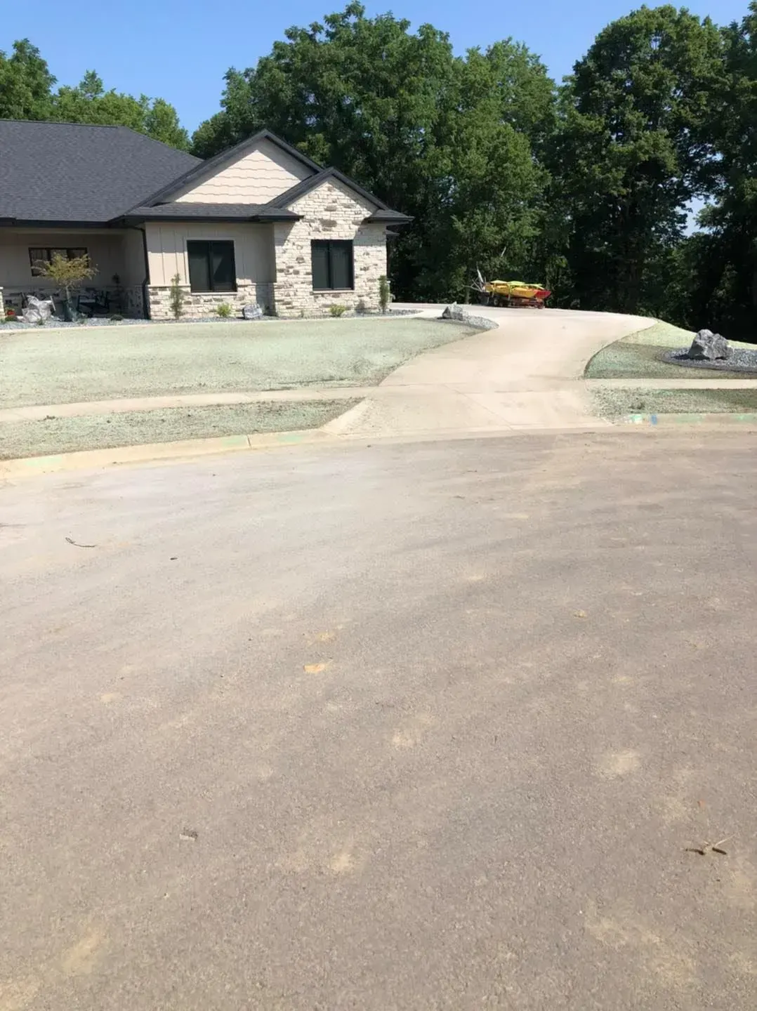 A house with stone and beige siding sits at the end of a long concrete driveway surrounded by trees and lawn.