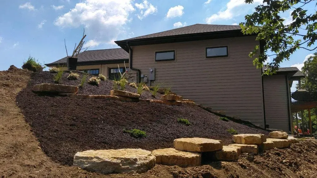A house with tan siding sits behind a sloped yard covered in dark mulch and accented with large natural stone steps.