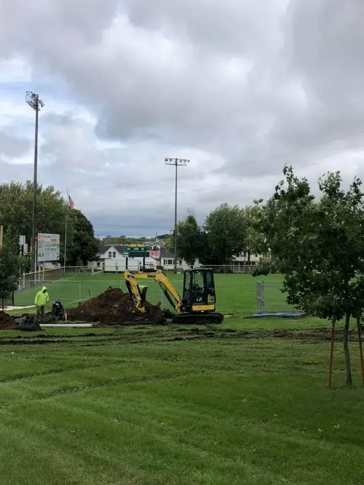 A yellow excavator sits in a grassy park, digging a trench near workers in neon safety gear under a cloudy sky.