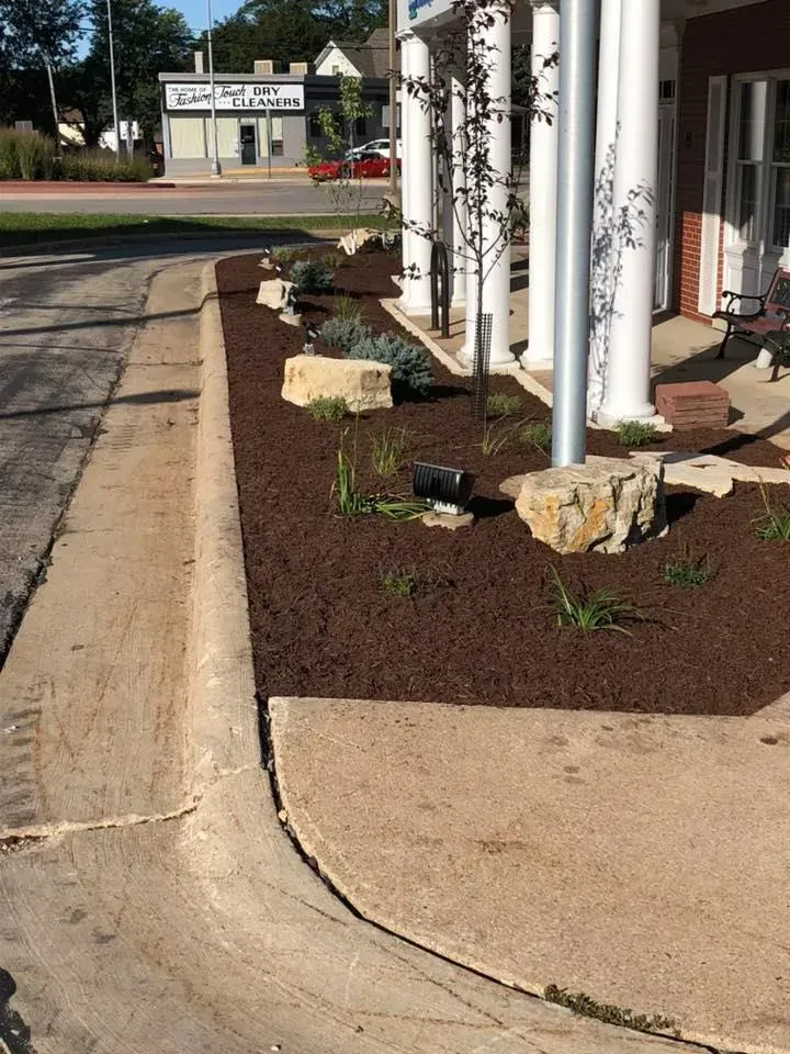 A landscape bed with brown mulch, decorative boulders, and small shrubs along the front of a building with white columns.