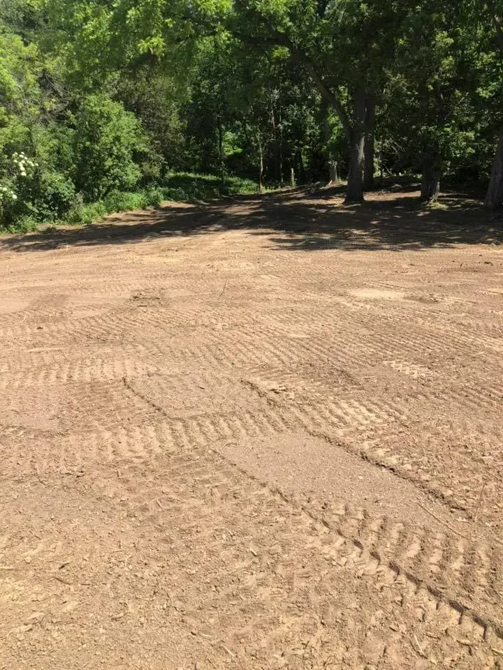 A cleared, dirt-covered lot with visible heavy equipment tire tracks, bordered by a dense line of green trees.