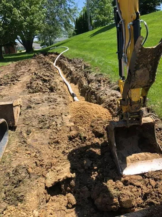 A yellow excavator sits in a grassy yard next to a trench containing a section of white drainage pipe and gravel.