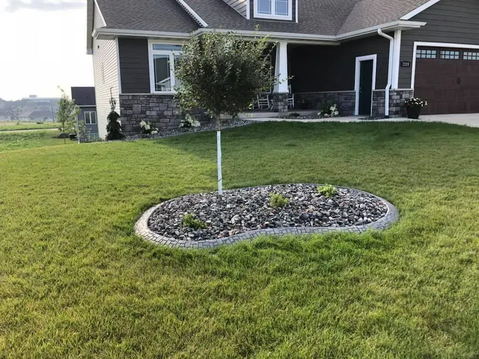 A suburban house with a brown garage and stone siding features a circular rock garden with a small tree in the front lawn.