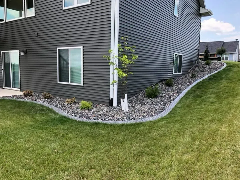 A dark gray house exterior with a curved landscaping bed featuring gray stones, mulch, small plants, and a downspout.