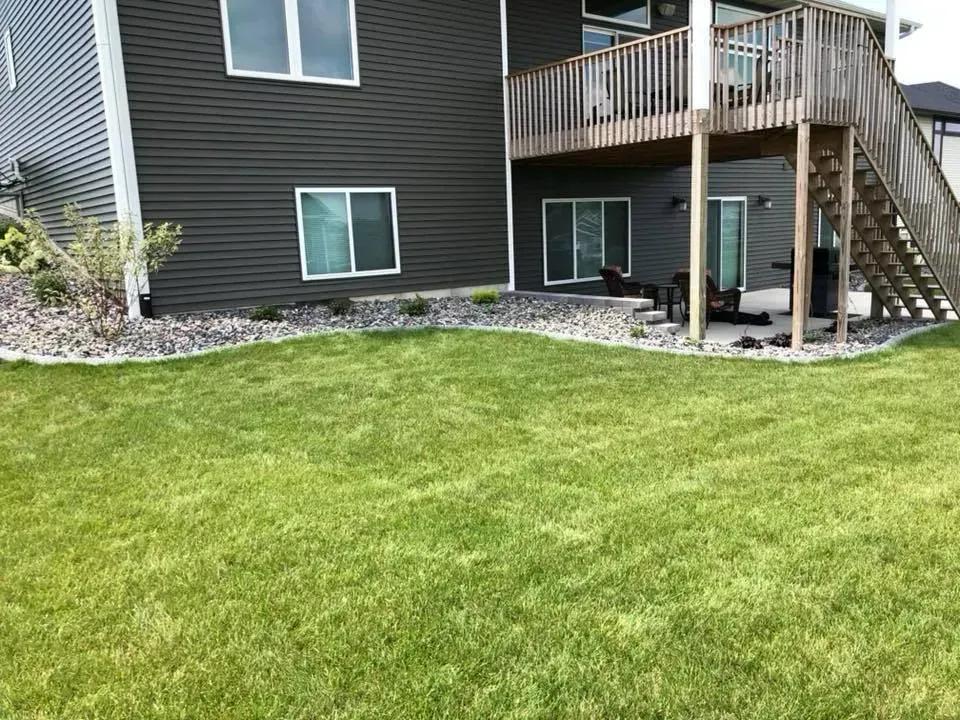 A dark-sided residential house with a lawn, landscaping rocks, and a raised wooden deck with stairs in the backyard.