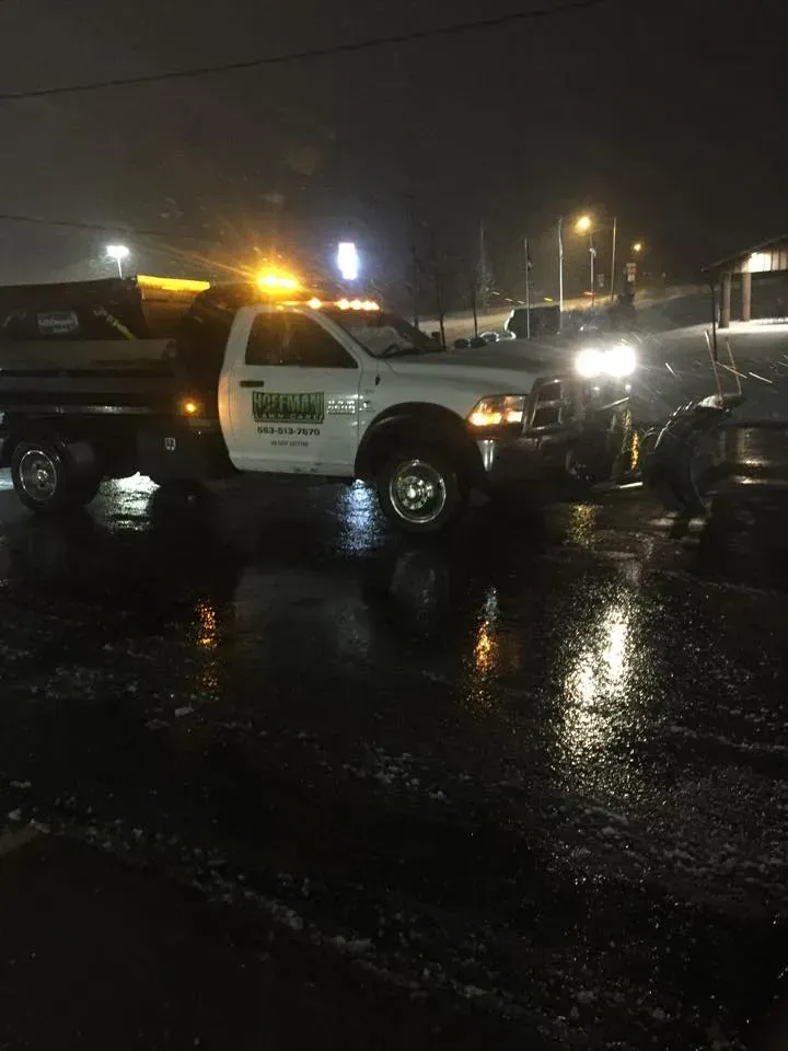 A white snowplow truck with a green logo is parked on a wet, dark surface at night under streetlights.