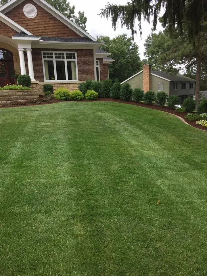 A well-manicured lawn leads to a brick and cedar home, with a row of green shrubs and a glimpse of a house in the background.