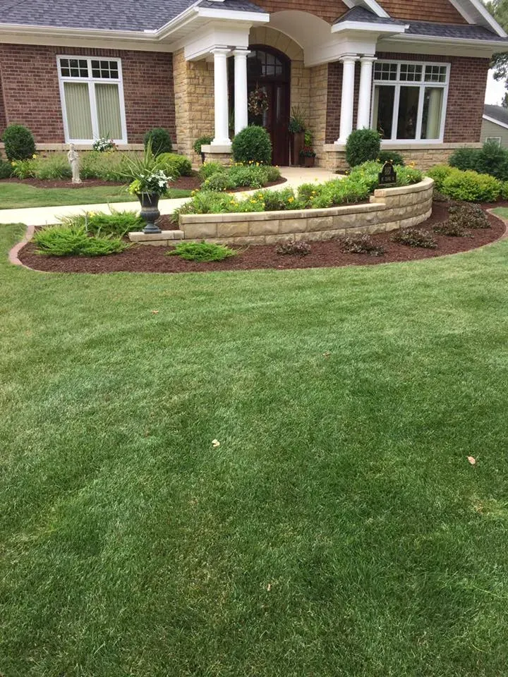 A tan stone retaining wall creates a curved flower bed in front of a brick house with white columns and green landscaping.