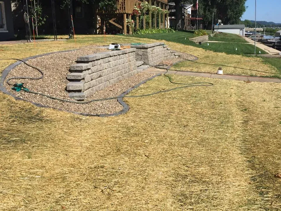 A tiered stone retaining wall set in a gravel bed within a grassy residential lawn on a sunny day.