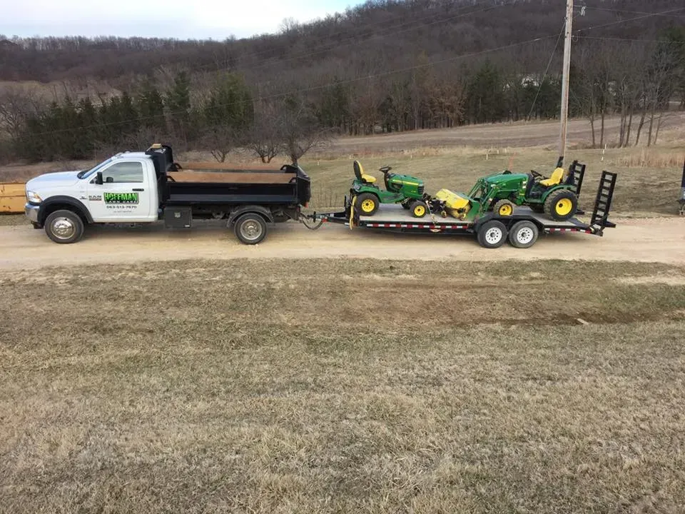 A white work truck pulling a flatbed trailer loaded with two green John Deere tractors on a dirt road.