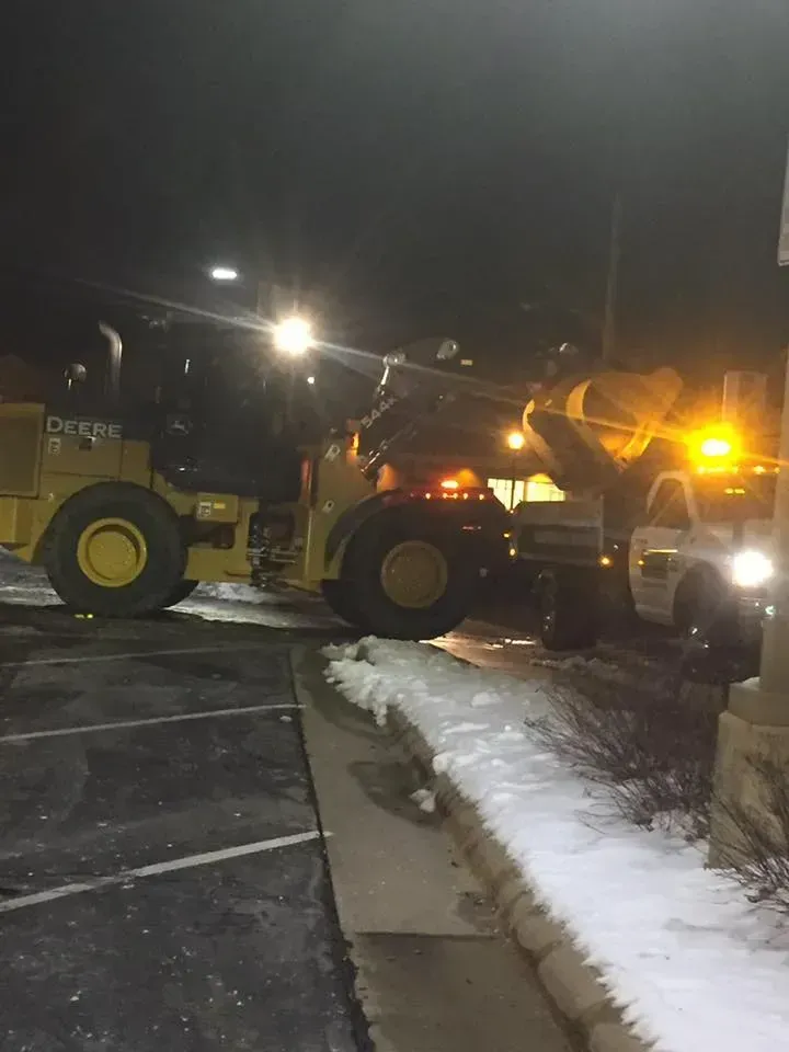A yellow John Deere wheel loader dumping snow into the back of a white dump truck at night in a parking lot.