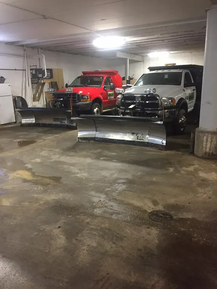 Two trucks with snowplows parked inside a dimly lit garage.