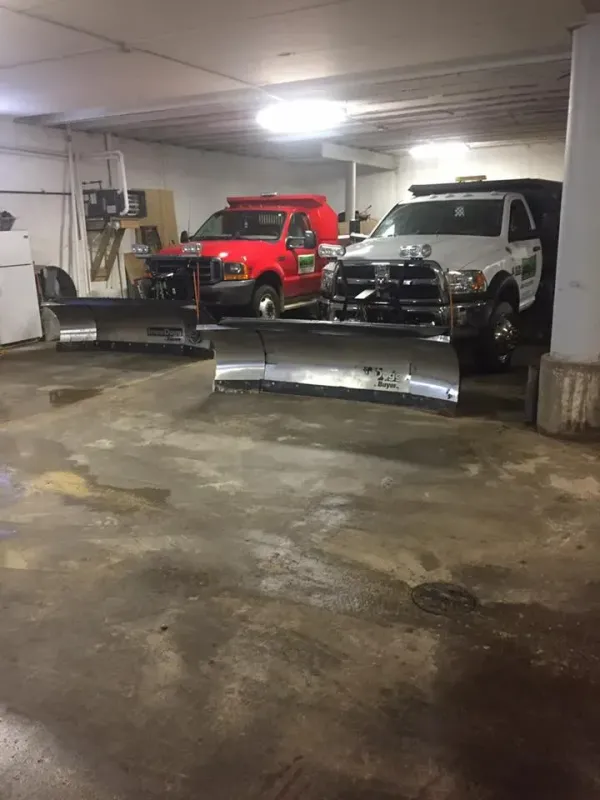 Two heavy-duty trucks with snowplows attached are parked side-by-side inside a dimly lit indoor storage garage.