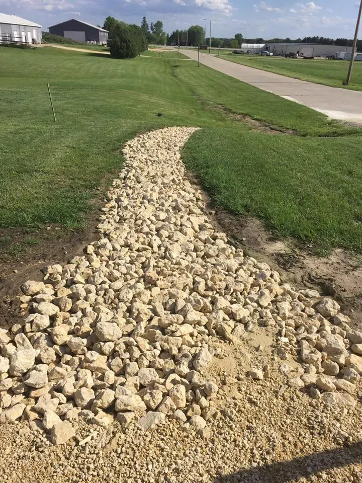 A light-colored stone drainage swale curves through a green grass lawn leading toward a paved road in a rural setting.