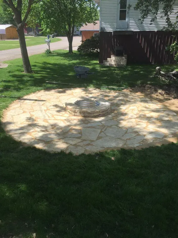 A circular stone patio with a central stone fire pit in a residential backyard, surrounded by a lush green lawn and trees.