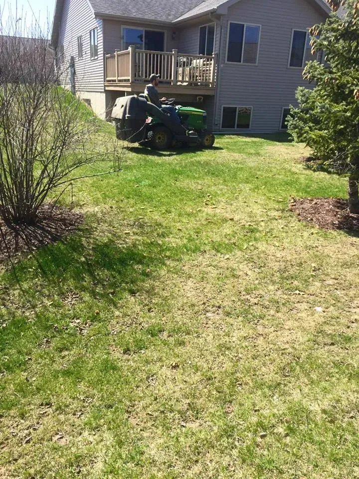 A person operates a green riding lawn mower on a grassy lawn in the backyard of a house.