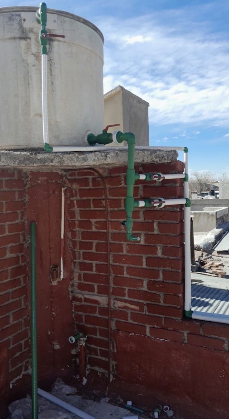 Una tubería verde sobre una pared de ladrillo rojo en la azotea, junto a un gran tanque beige bajo un cielo azul.