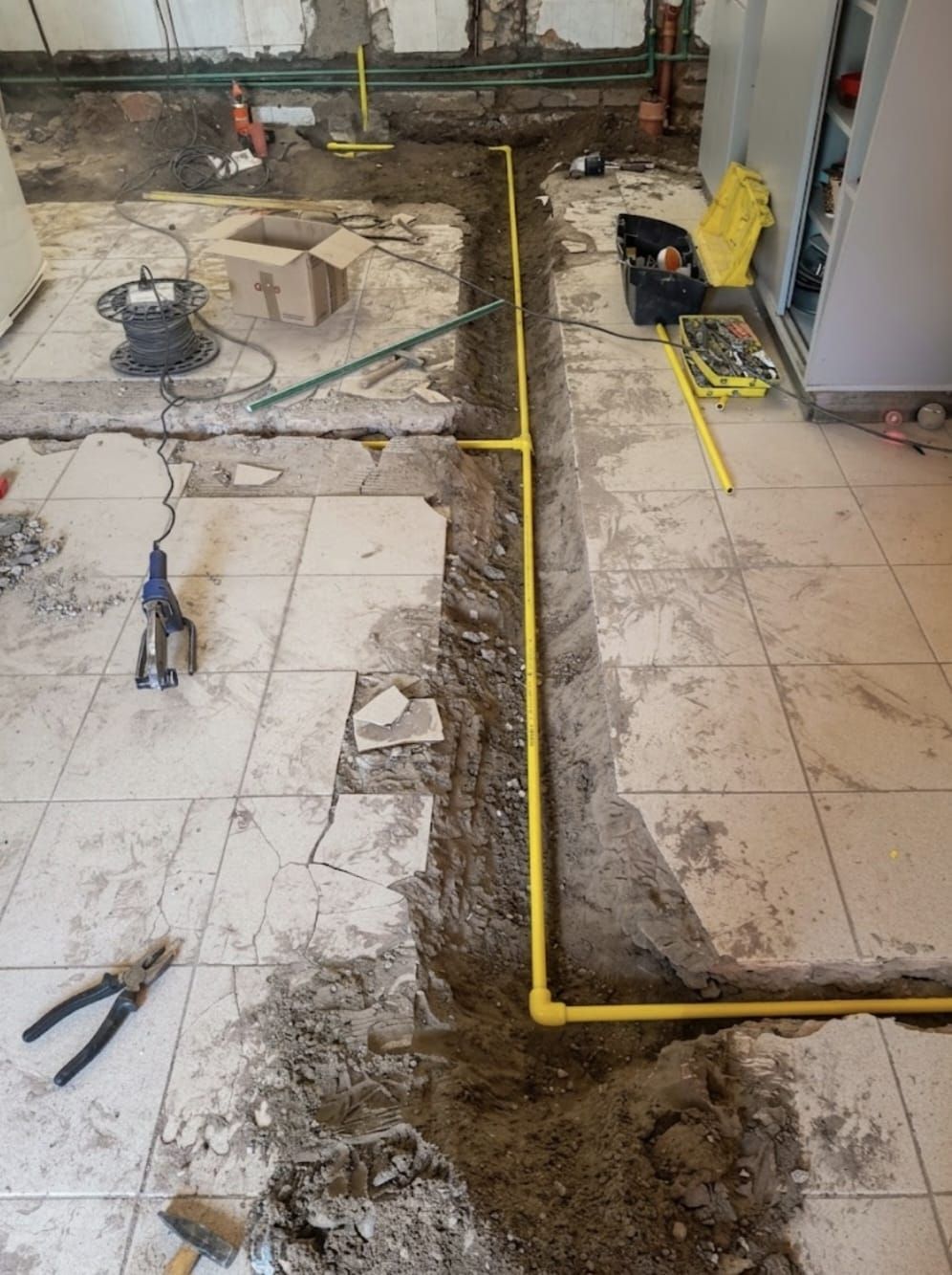 Piso de baldosas en renovación con zanja de tierra expuesta, herramientas y tuberías amarillas en un área de trabajo.