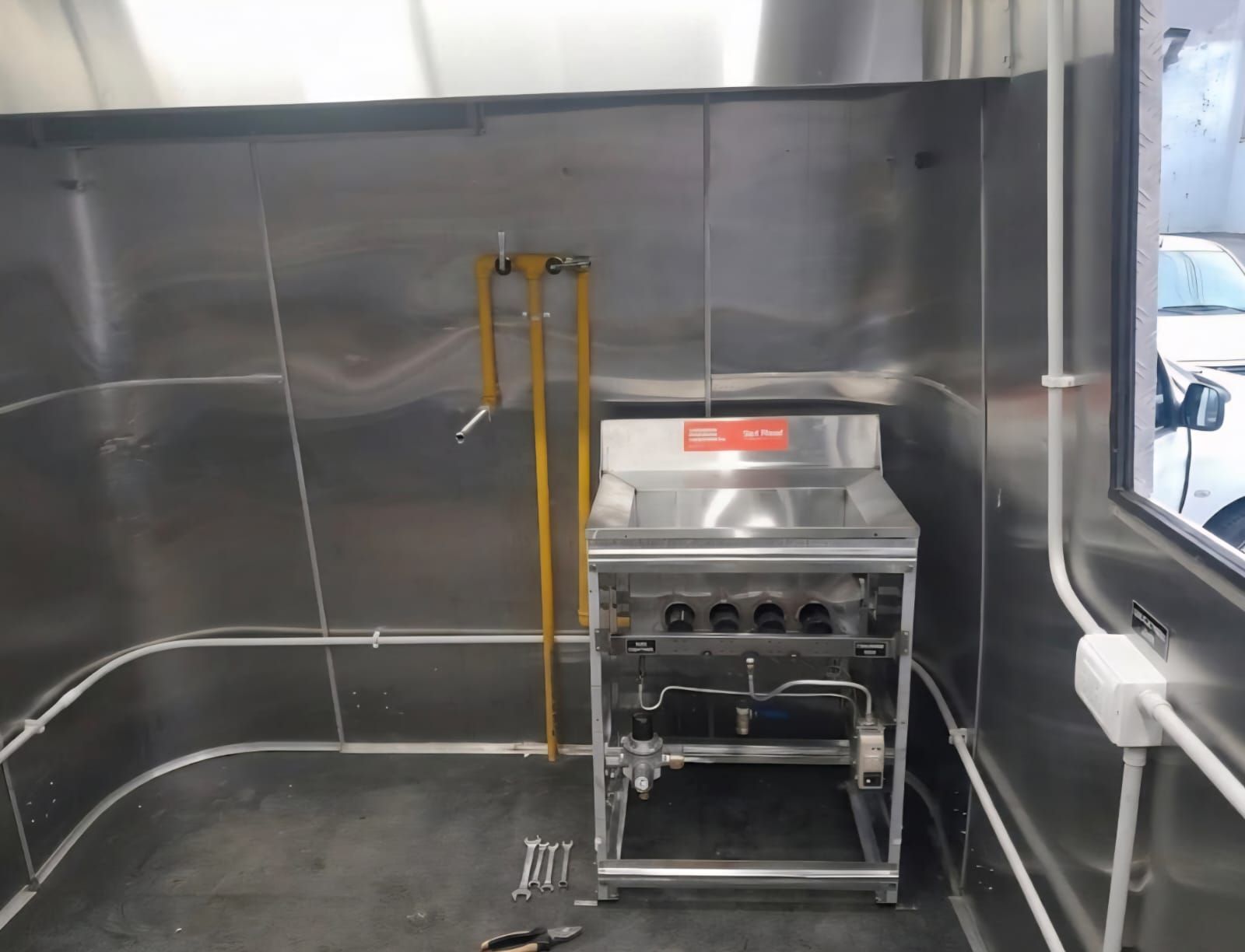 Zona de cocina de acero inoxidable con una parrilla compacta y tuberías de gas amarillas contra una pared metálica.