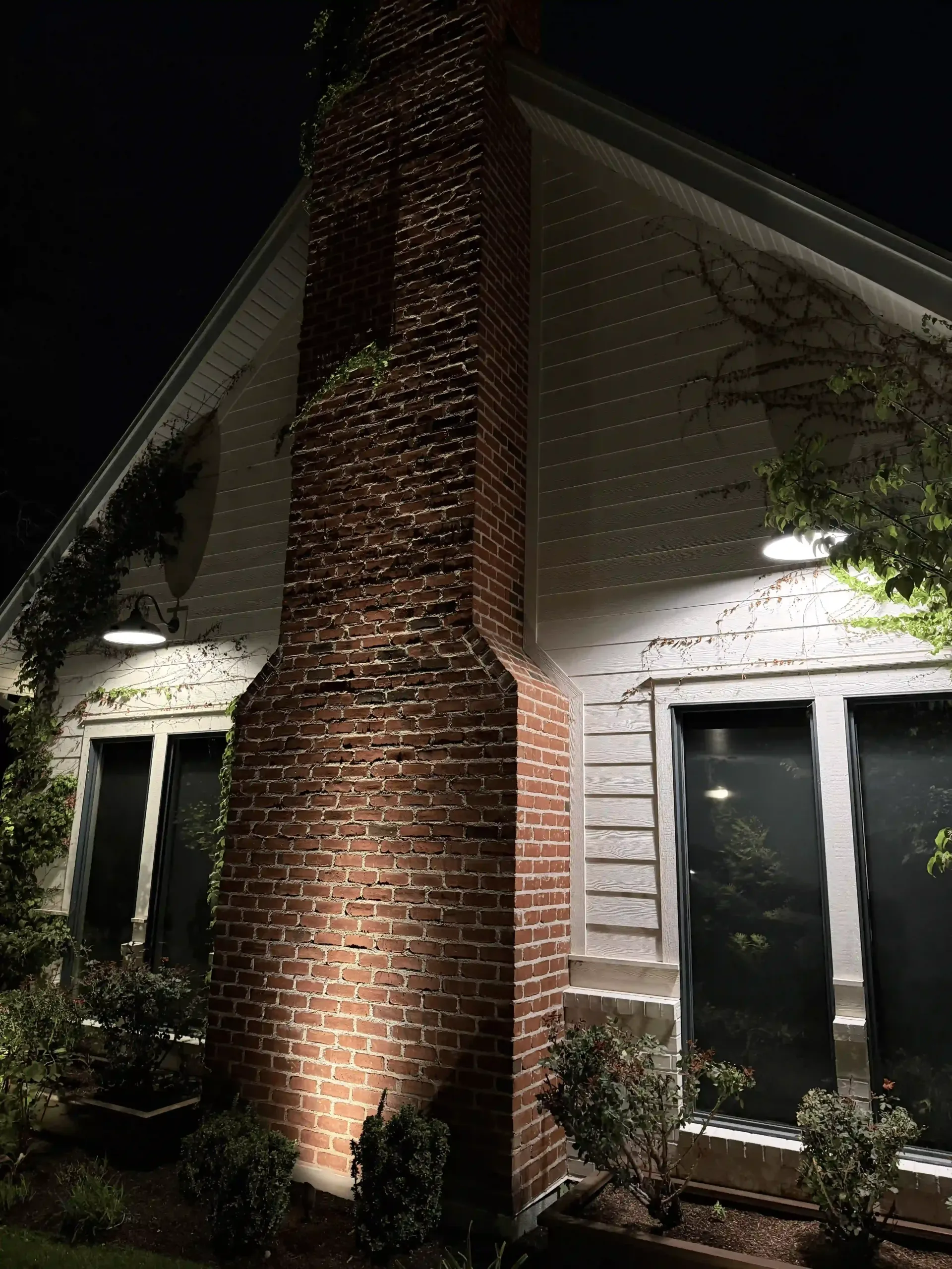 Brick chimney on a white house at night, lit by exterior lights, with dark windows and shrubs below. Brick chimney on a white house at night, lit by exterior lights, with dark windows and shrubs below.
