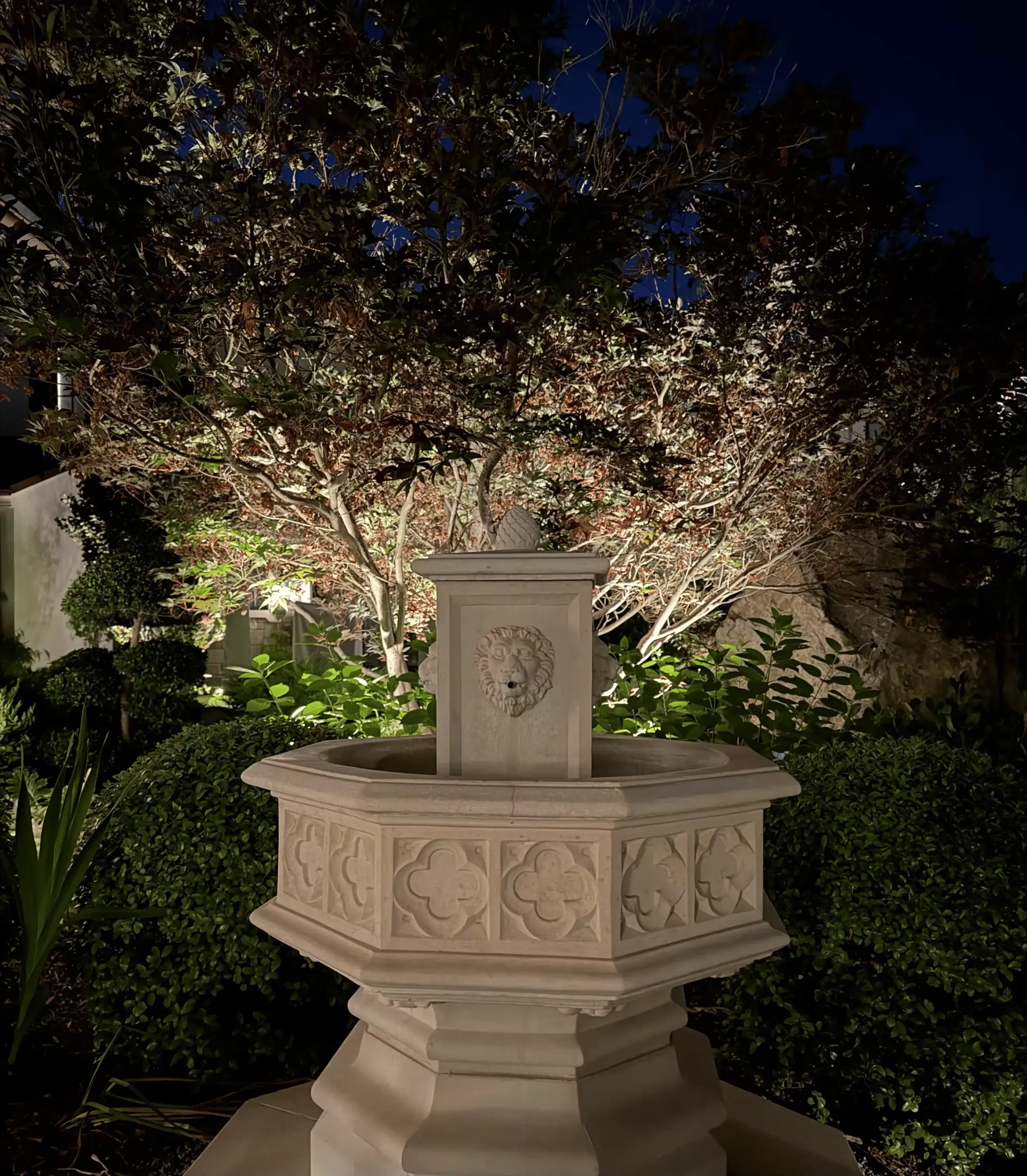 Illuminated stone fountain in a garden at night with trees and bushes behind it