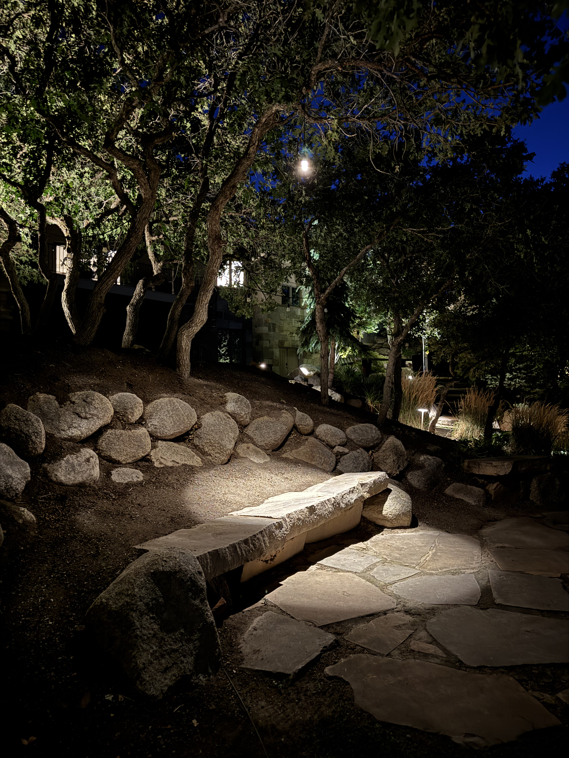 A wet stone tile path reflecting warm landscape lights in the rain, leading to a modern house with stone siding at night.